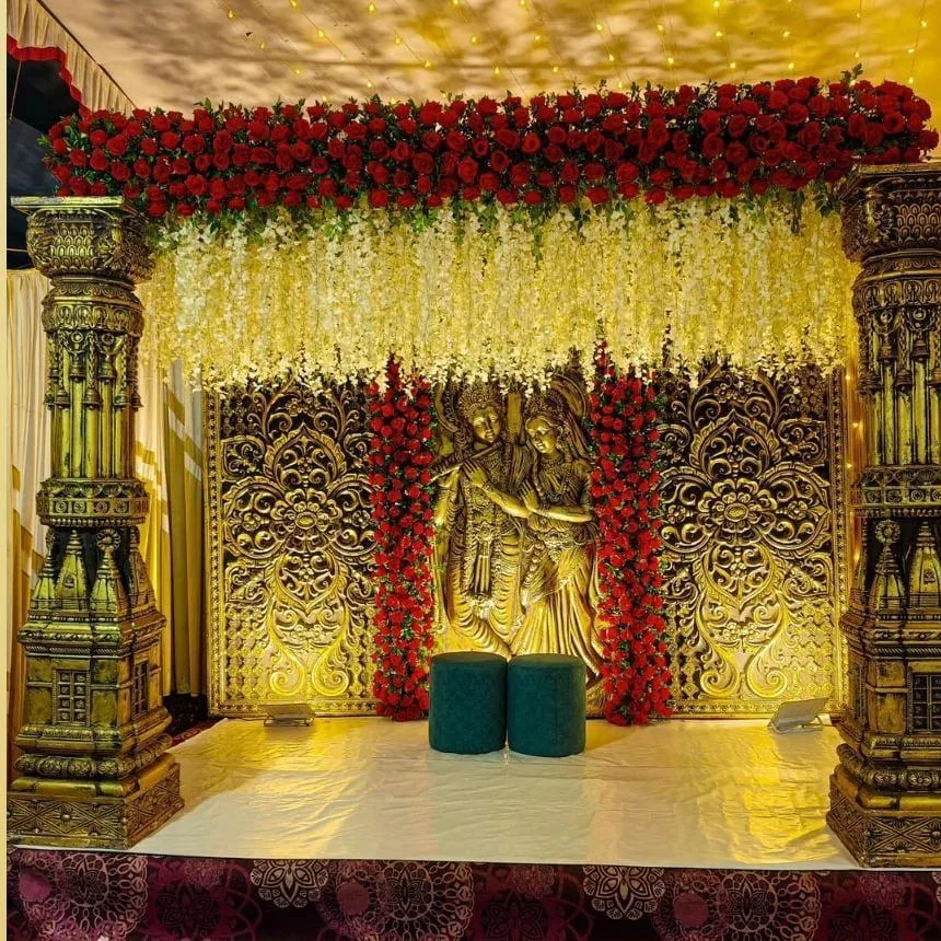 Golden carved stage with a Radha-Krishna backdrop and red rose garlands